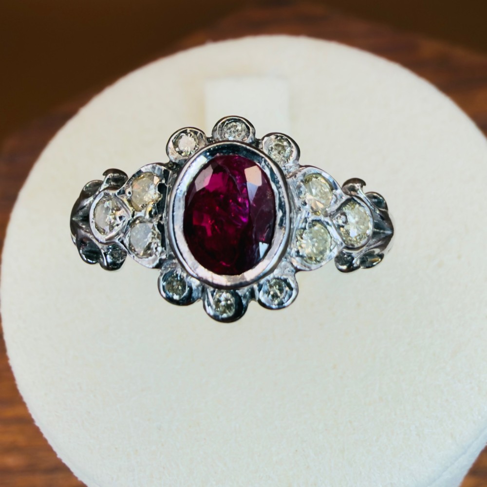 An 18ct white gold cluster ring featuring a central oval bezel-set red ruby framed by small round diamonds and decorative shoulders.