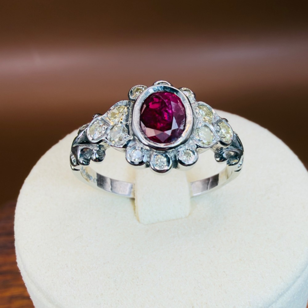 An 18ct white gold cluster ring featuring a central oval bezel-set red ruby framed by small round diamonds and decorative shoulders. View IX