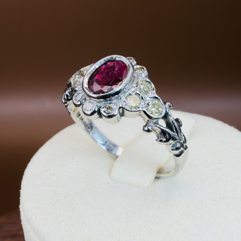 An 18ct white gold cluster ring featuring a central oval bezel-set red ruby framed by small round diamonds and decorative shoulders. View X