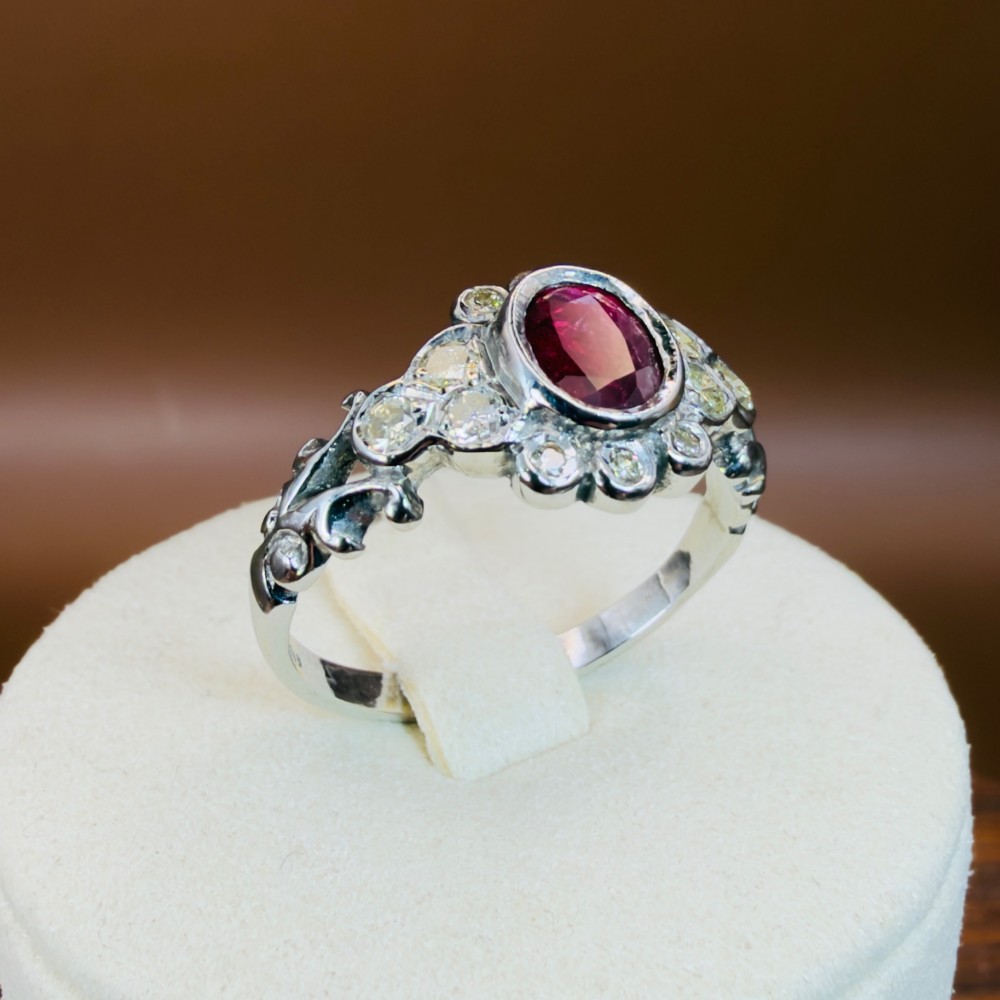An 18ct white gold cluster ring featuring a central oval bezel-set red ruby framed by small round diamonds and decorative shoulders. View XI