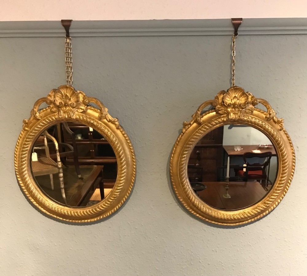 A pair of circular wall mirrors featuring carved gilt frames with top shell pediments, rope-twist inner borders, and chain suspensions.