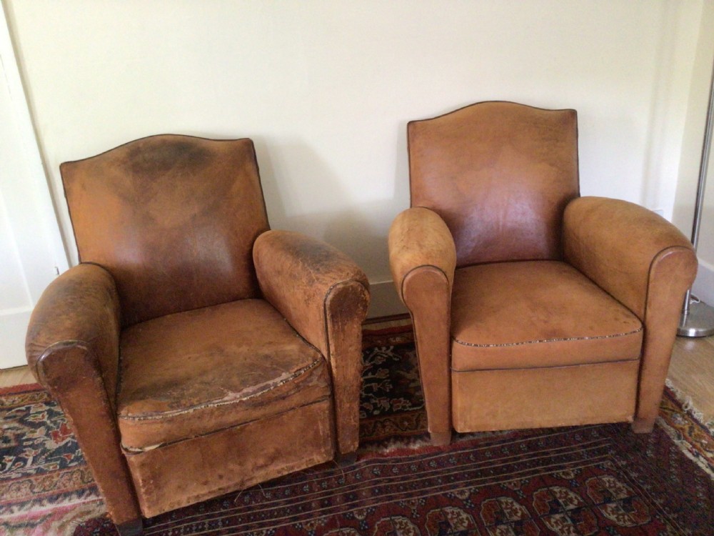 A pair of Art Deco tan leather club armchairs featuring rolled armrests, arched backrests, and aged surface patina.