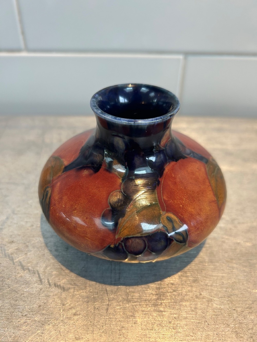A small squat ceramic vase featuring hand-painted pomegranate and berry motifs in shades of terracotta, purple, and green against a deep blue glazed background.