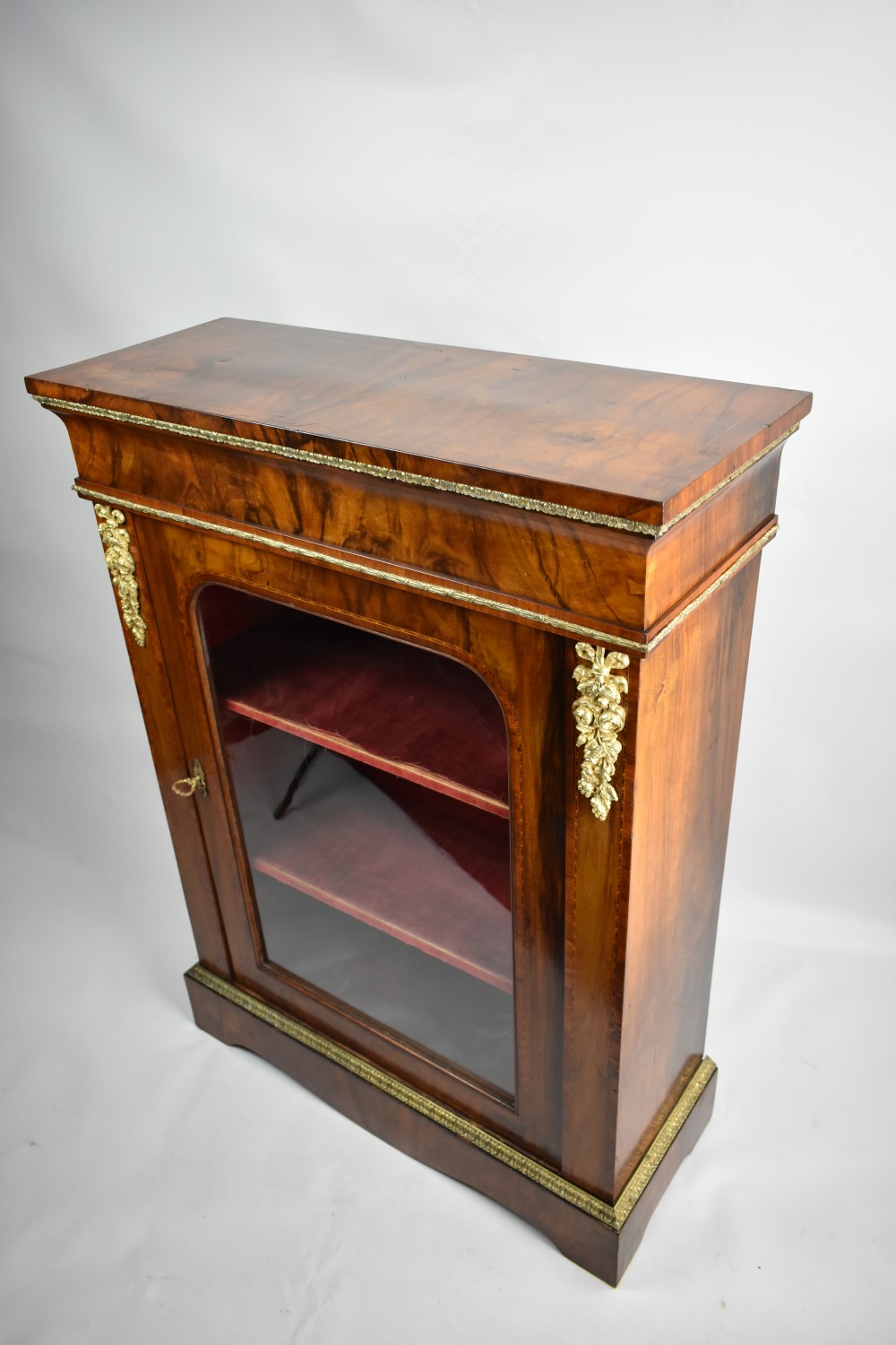 Mid-Victorian figured walnut pier cabinet featuring tulipwood banding, ormolu mounts, and a single glazed door showing velvet-lined shelves.