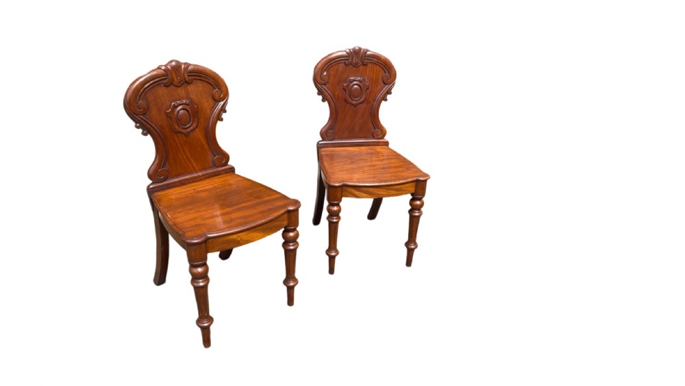 A pair of 19th-century William IV mahogany hall chairs featuring carved cartouche backrests, solid timber seats, and turned front legs. View IV