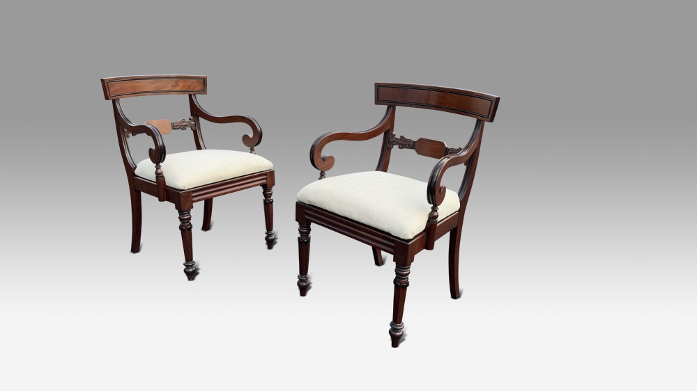 A pair of antique Regency mahogany armchairs featuring curved top rails with ebonised stringing, sweeping scroll armrests, cream upholstered seats, and turned front legs.