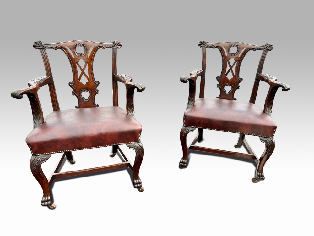 A pair of carved mahogany armchairs featuring openwork pierced splat backs, brown leather upholstered seats with brass studding, and cabriole legs ending in claw and ball feet.