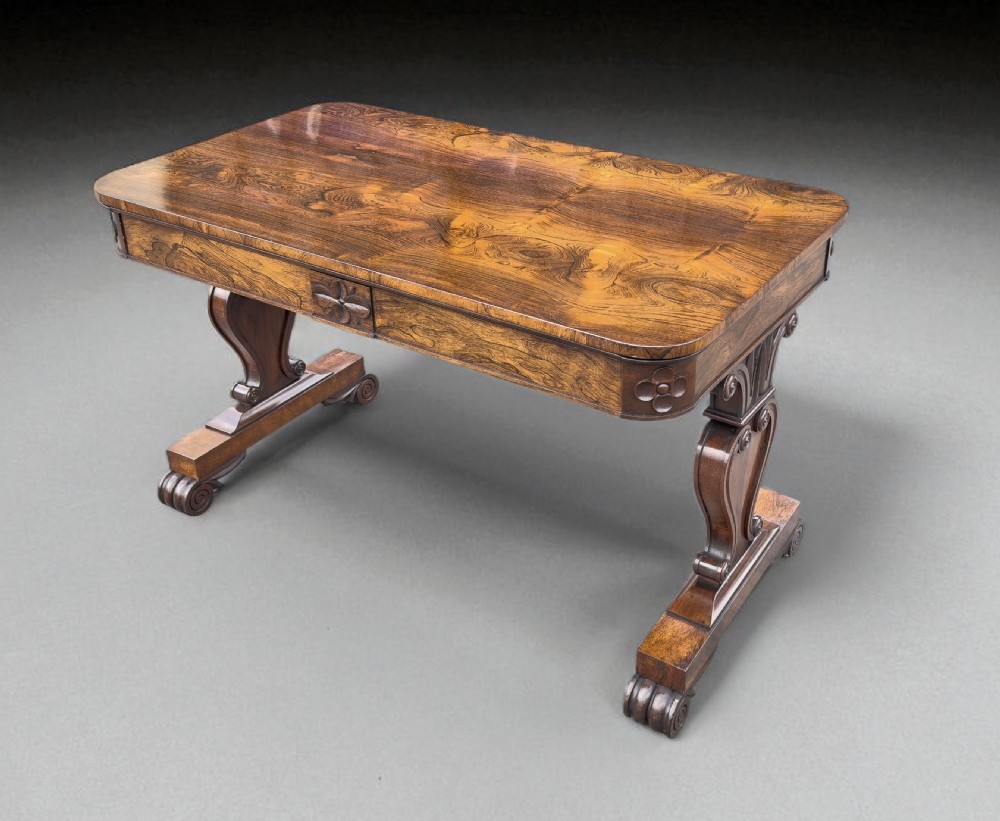 A Regency rosewood library table featuring a highly figured rectangular top, frieze drawer with carved corner patera, and scroll trestle supports.