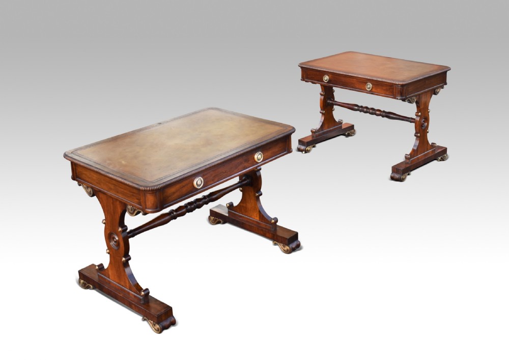 A pair of Regency rosewood writing tables featuring inset leather tops, single drawers with brass mounts, turned stretchers, and trestle end supports on castors.