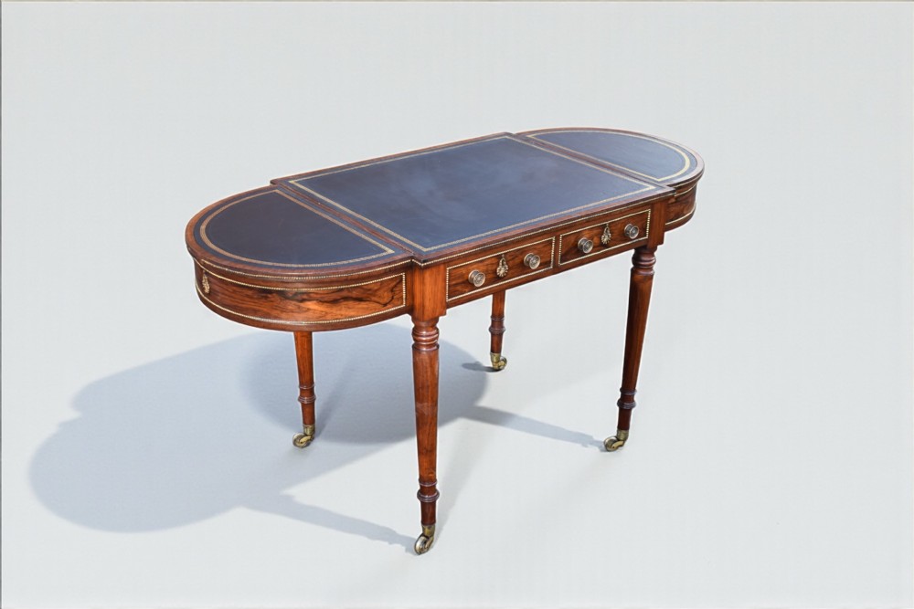 A Regency rosewood writing table featuring a dark leather inset top with brass beaded borders, frieze drawers, rounded ends, and turned legs on brass castors. View IV