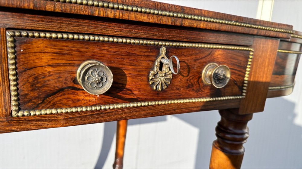 A Regency rosewood writing table featuring a dark leather inset top with brass beaded borders, frieze drawers, rounded ends, and turned legs on brass castors. View VII