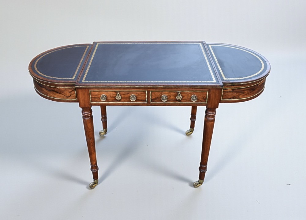 A Regency rosewood writing table featuring a dark leather inset top with brass beaded borders, frieze drawers, rounded ends, and turned legs on brass castors. View III