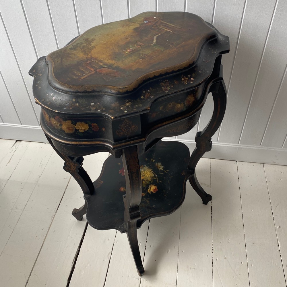 A Victorian ebonised black wooden sewing table featuring hand-painted floral designs, a painted landscape scene on the lid, cabriole legs, and a lower undertier.