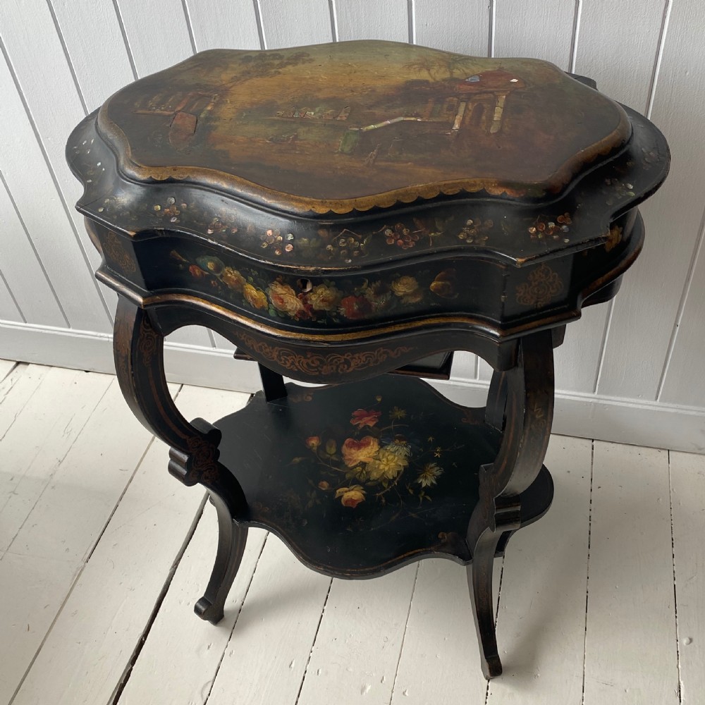 A Victorian ebonised black wooden sewing table featuring hand-painted floral designs, a painted landscape scene on the lid, cabriole legs, and a lower undertier. View VI