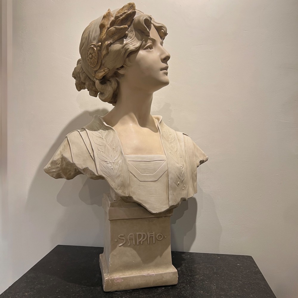 An Art Nouveau cream-toned sculptural bust of the classical poet Sappho wearing a golden laurel wreath, mounted on a plinth inscribed with her name.