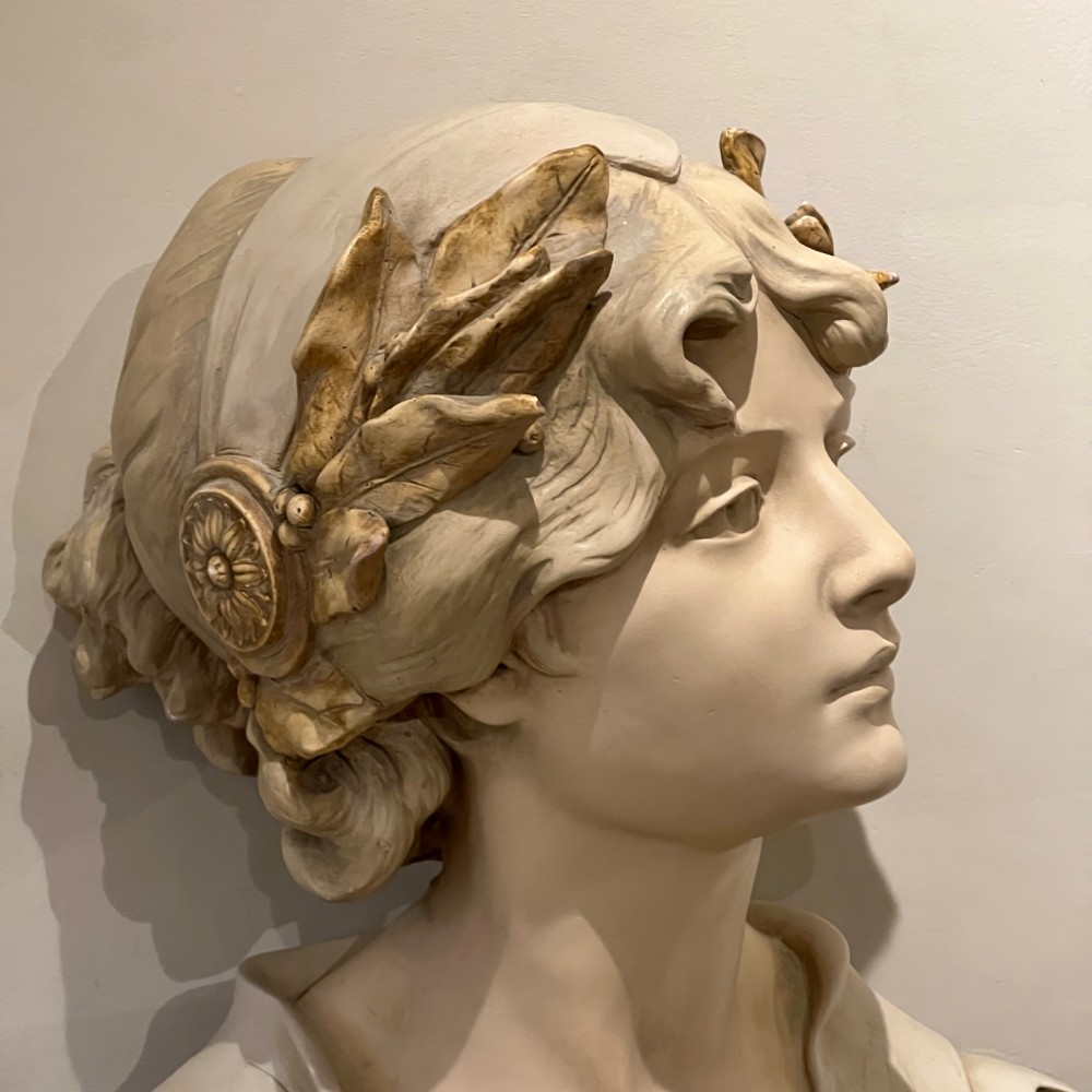 An Art Nouveau cream-toned sculptural bust of the classical poet Sappho wearing a golden laurel wreath, mounted on a plinth inscribed with her name. View VI