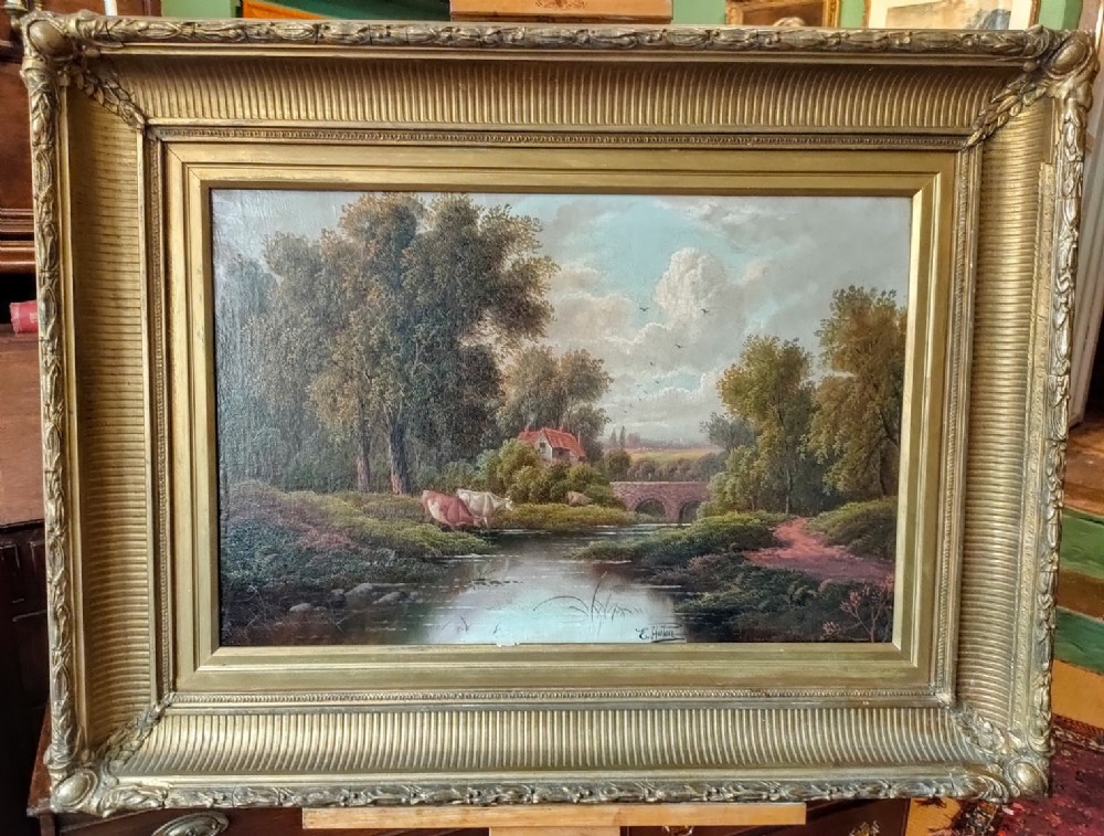An oil painting depicting cows grazing beside a river with a stone bridge and cottage in the background, set within an ornate gilded frame.