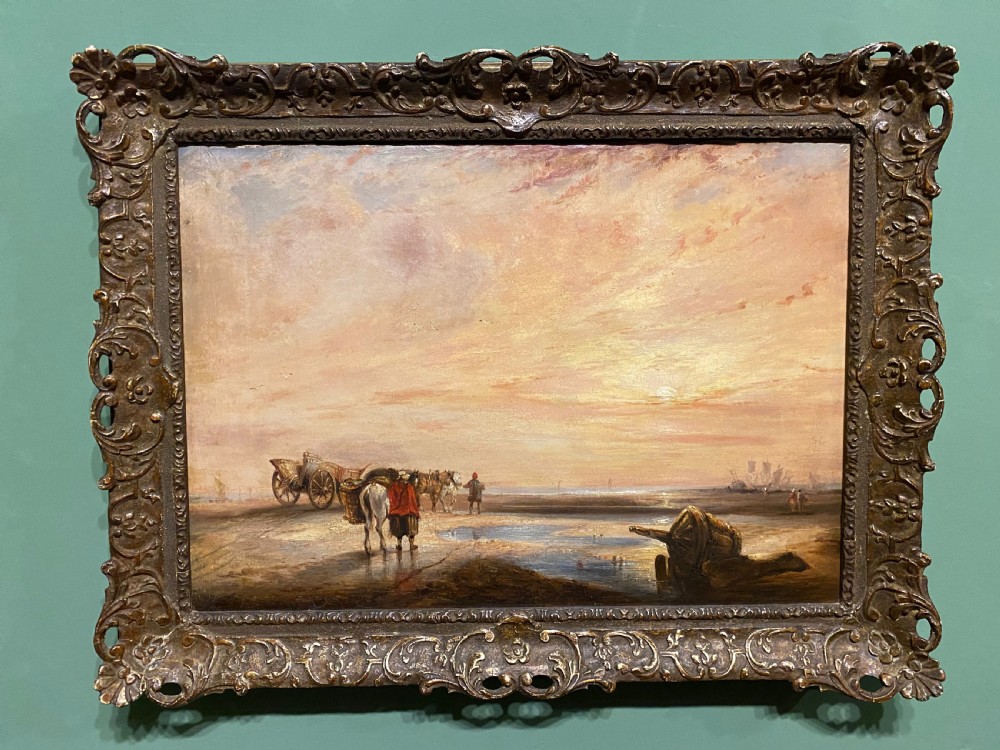 A nineteenth-century coastal oil painting depicting figures and horse-drawn carts on a shore at low tide during sunset, framed in an ornate gilded frame.