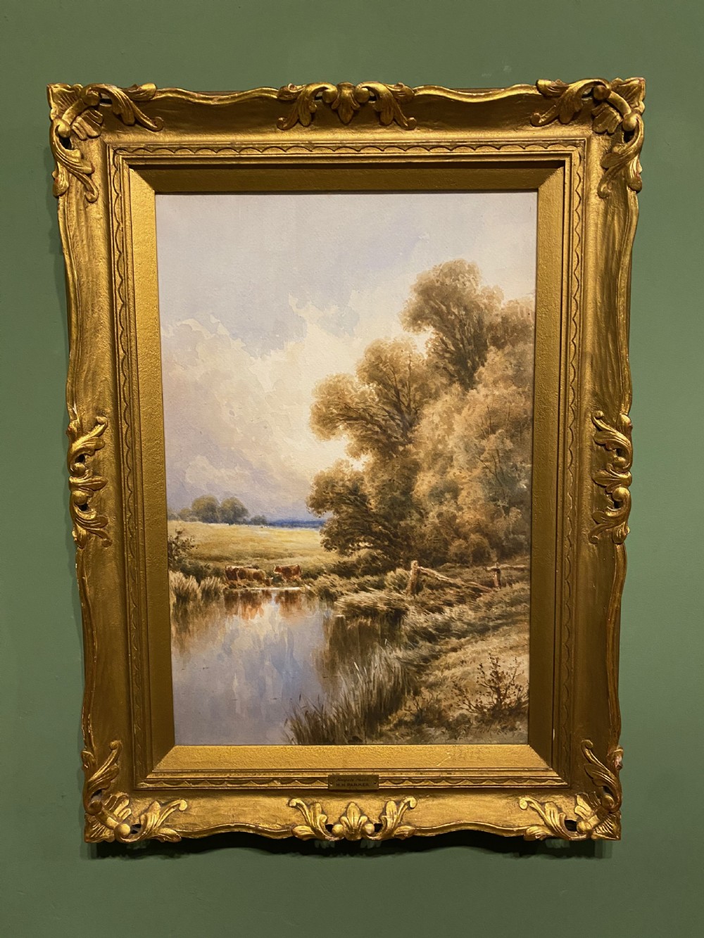 A 19th-century framed watercolour landscape painting depicting cattle resting by a river bank alongside trees, enclosed within a gold swept frame.