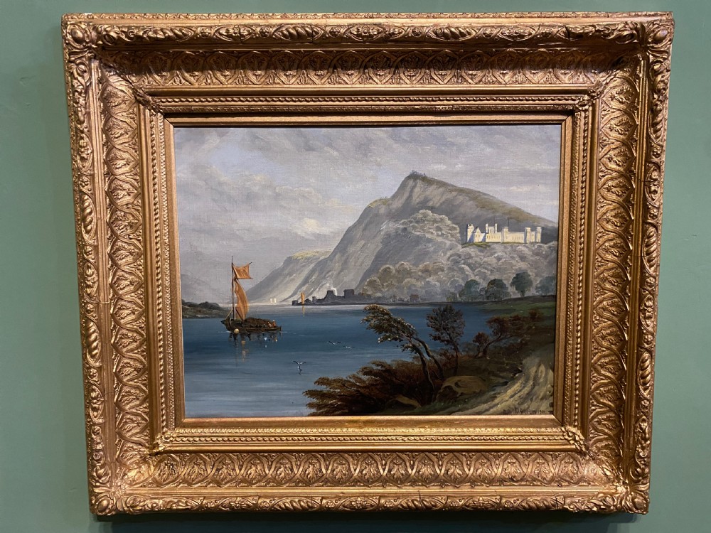 An antique oil painting depicting a river scene with a sailboat, distant castle on a hill, and mountains, housed in an ornate giltwood frame.