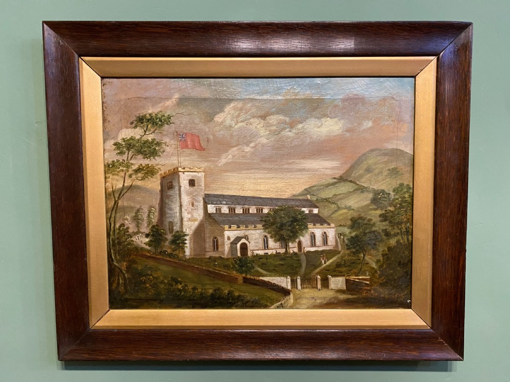 A 19th-century oil painting depicting a parish church flying a red flag set within a hilly countryside landscape, housed in a wide wooden frame with a gold inner border.