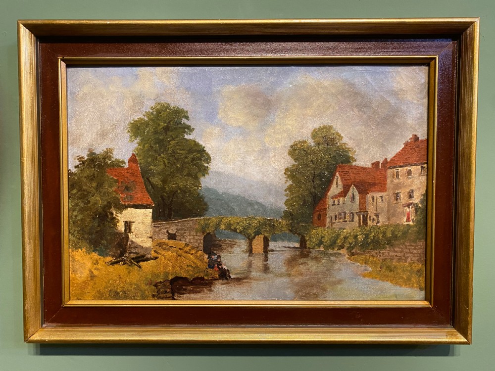 A 19th-century oil painting on canvas depicting a rural river scene with a stone bridge, village cottages, and seated figures on a grassy bank, presented in a dark wooden frame with gilt trim.