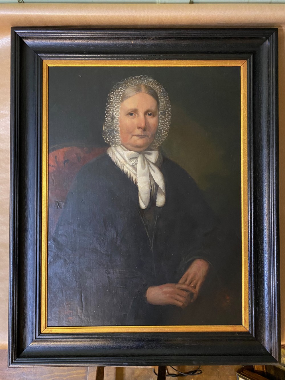A 19th-century oil portrait depicting a seated woman wearing a lace bonnet and white neckerchief, contained in a black wooden frame with a gold inner slip.