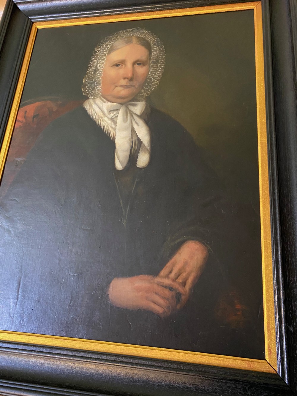 A 19th-century oil portrait depicting a seated woman wearing a lace bonnet and white neckerchief, contained in a black wooden frame with a gold inner slip. View III