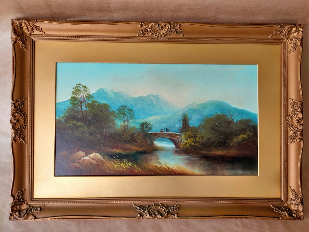 An antique landscape oil painting portraying two figures standing on a stone arch bridge over a river with blue mountains in the background, set inside an ornate gold frame.