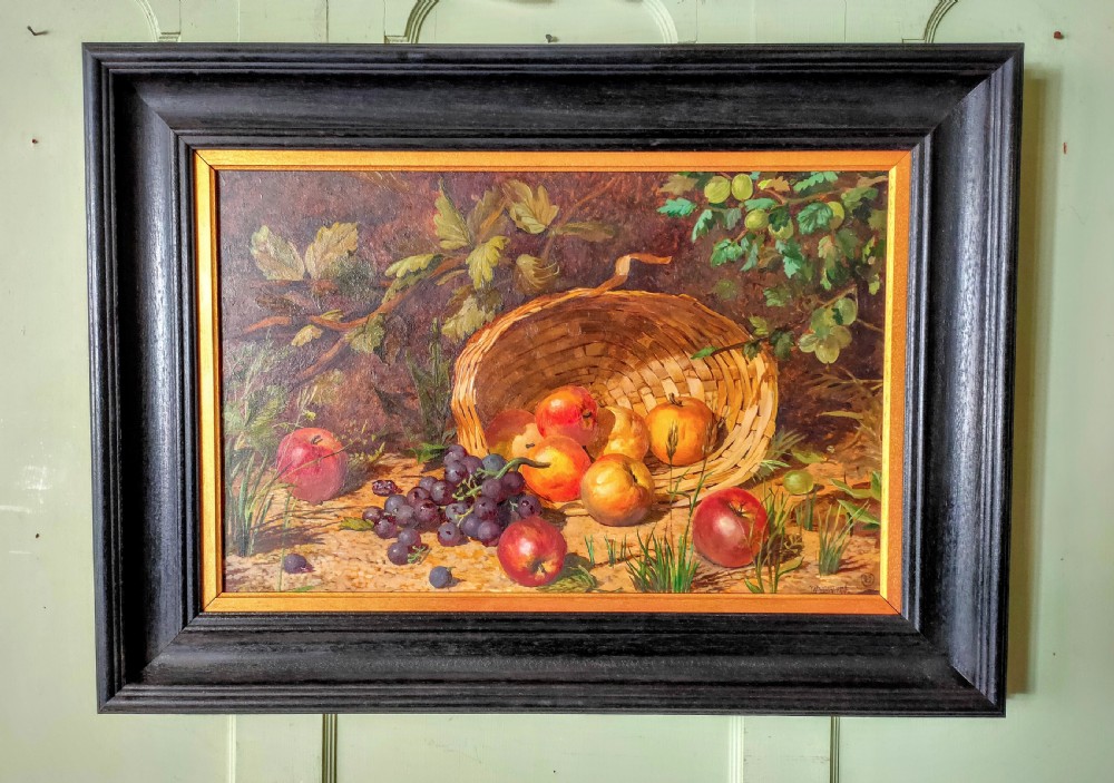 A framed oil painting featuring a tilted wicker basket with apples and purple grapes resting on ground surrounded by foliage.