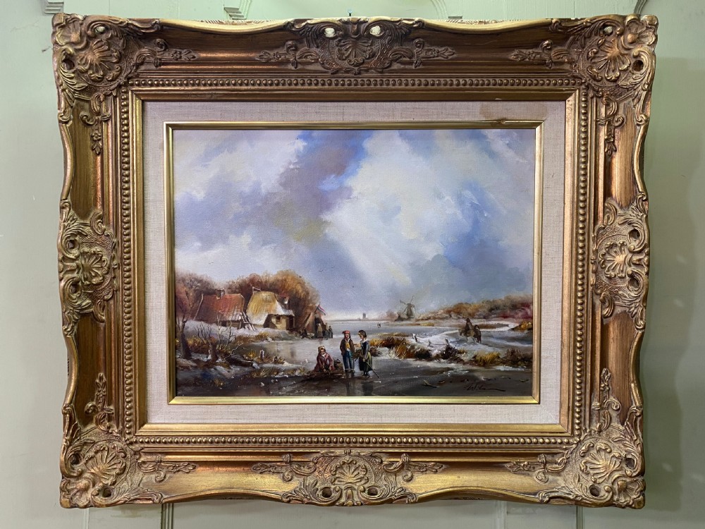 An oil painting depicting a winter landscape with figures near a thatched cottage on a frozen river, set in an ornate giltwood frame.