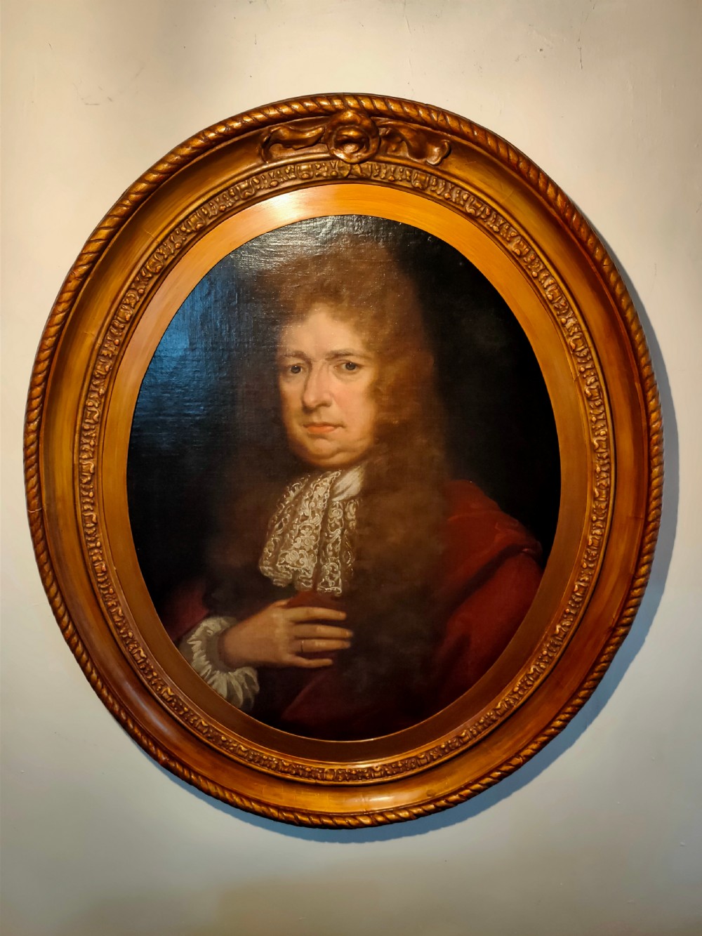 A seventeenth-century oval oil painting on canvas depicting a gentleman with a long wig, lace cravat, and crimson cloak, set within a carved wood frame.