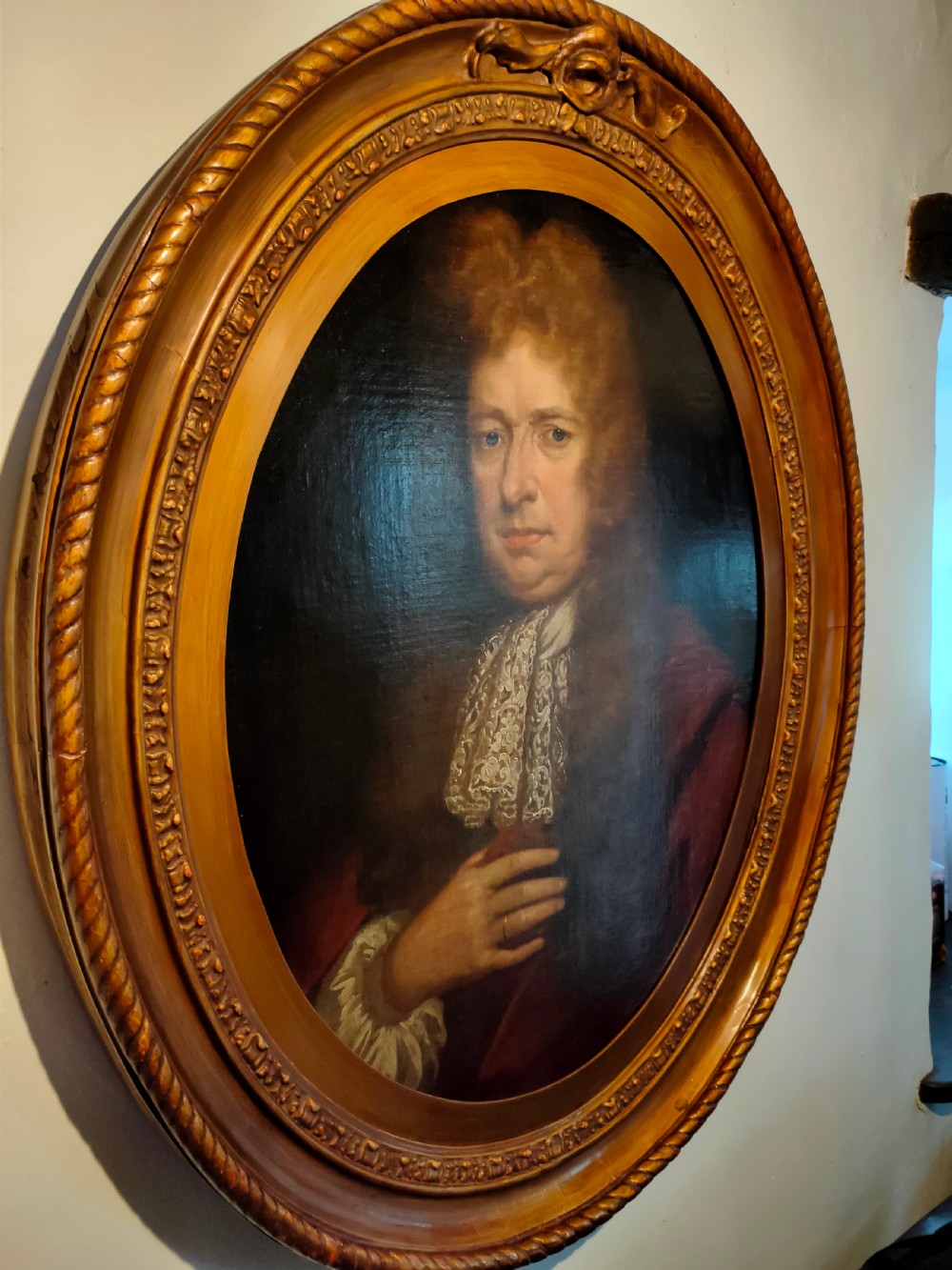 A seventeenth-century oval oil painting on canvas depicting a gentleman with a long wig, lace cravat, and crimson cloak, set within a carved wood frame. View II