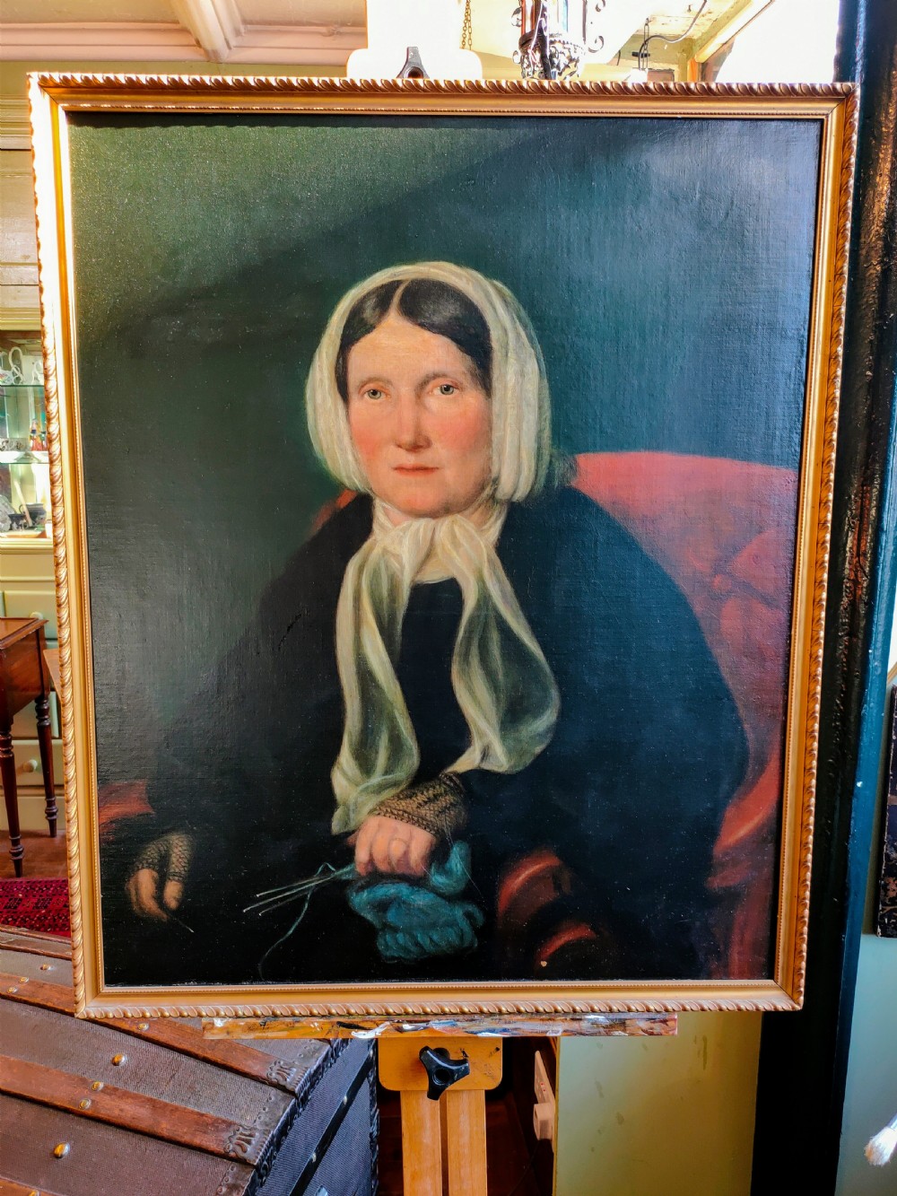A 19th-century oil painting depicting a seated woman in dark attire and a white headscarf holding knitting needles, presented in a gold frame.
