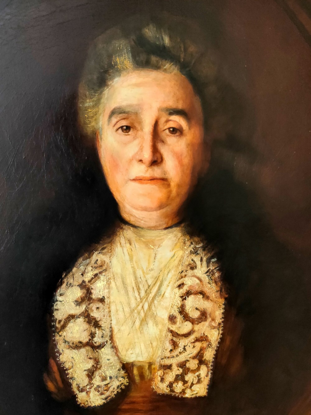 An antique oil painting depicting an elder woman in a lace stole set against a dark background within a wide gilt frame. View II