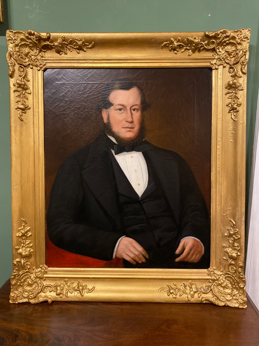 A 19th-century oil painting on canvas depicting a seated gentleman in black formal wear, set within an ornate gilded frame.