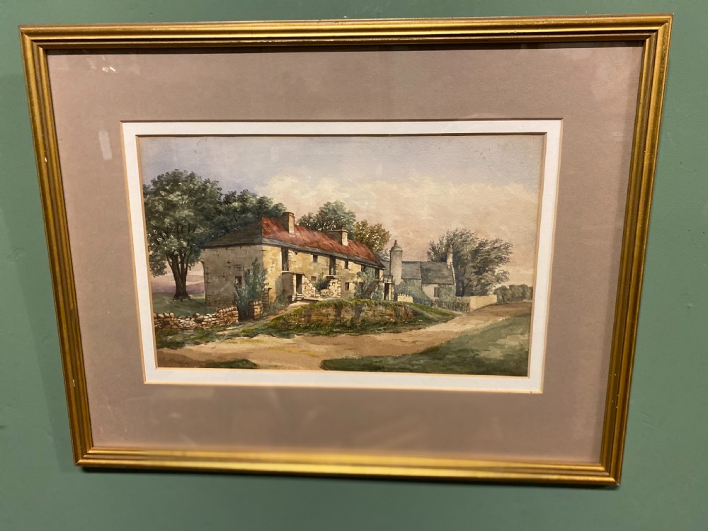 A framed watercolour painting depicting stone cottages and countryside terrain, displayed within a wide mount and slender gilt frame.