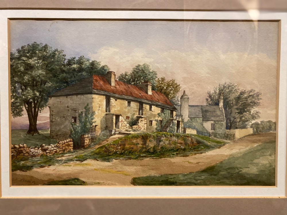A framed watercolour painting depicting stone cottages and countryside terrain, displayed within a wide mount and slender gilt frame. View II