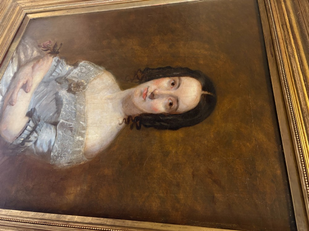 A mid-19th-century oil painting on canvas depicting a seated lady in a light blue dress with a lace collar, set inside a heavy gilded rectangular frame. View VI