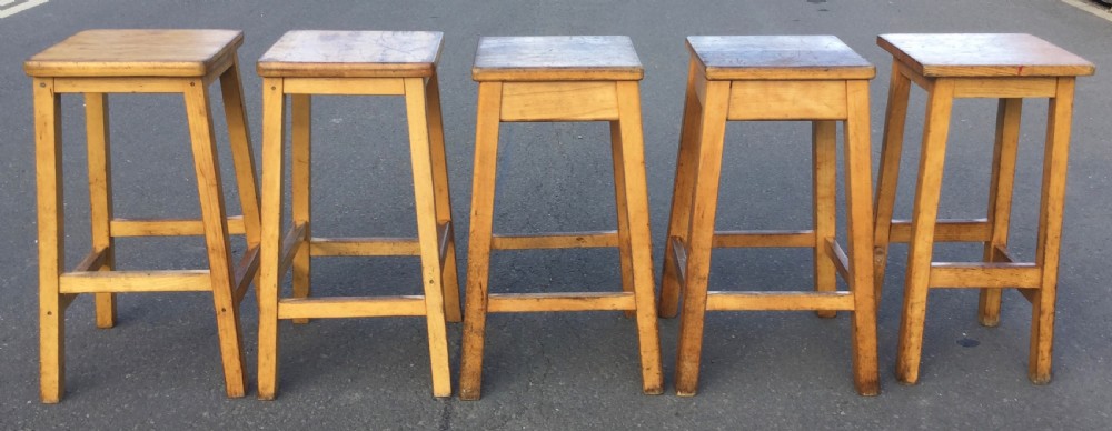 A row of five vintage wooden high stools featuring square flat seats, splayed legs, and four-sided footrest stretchers.