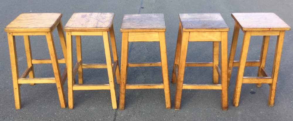 A row of five vintage wooden high stools featuring square flat seats, splayed legs, and four-sided footrest stretchers. View II