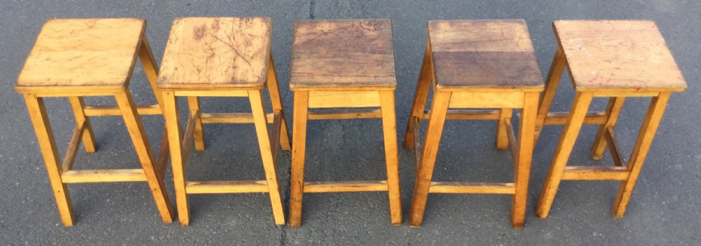 A row of five vintage wooden high stools featuring square flat seats, splayed legs, and four-sided footrest stretchers. View V