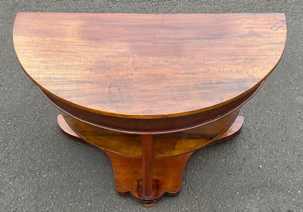 A Victorian mahogany demi-lune console table featuring a semicircular top, an intermediate tier shelf, and a shaped plinth base. View VI