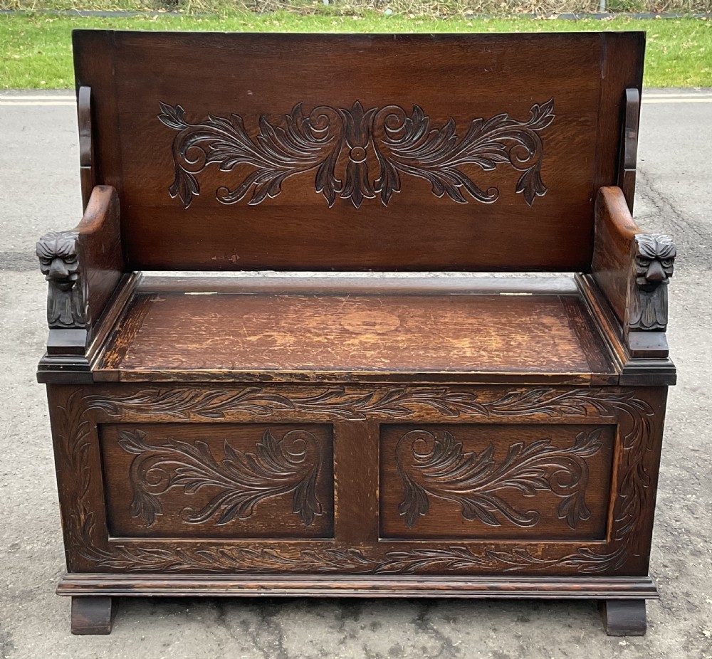 A dark oak monks bench featuring foliate scroll carving on the backrest and base panels, carved lion head armrests, and a hinged lift-up seat.
