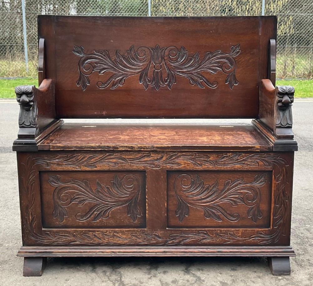 A dark oak monks bench featuring foliate scroll carving on the backrest and base panels, carved lion head armrests, and a hinged lift-up seat. View II