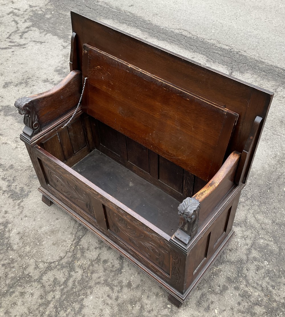 A dark oak monks bench featuring foliate scroll carving on the backrest and base panels, carved lion head armrests, and a hinged lift-up seat. View VII
