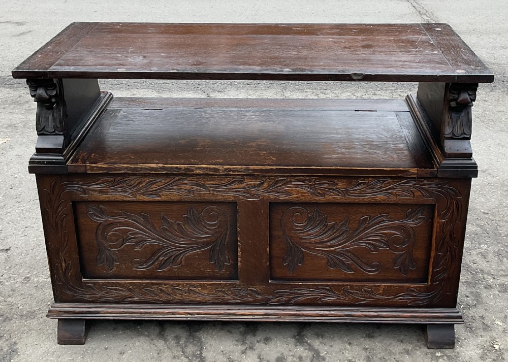 A dark oak monks bench featuring foliate scroll carving on the backrest and base panels, carved lion head armrests, and a hinged lift-up seat. View IX