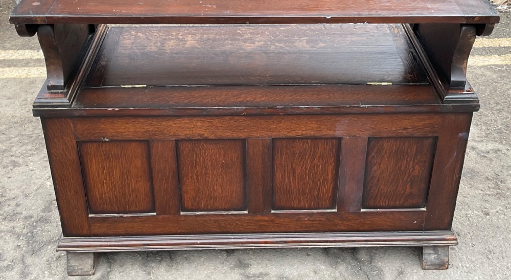 A dark oak monks bench featuring foliate scroll carving on the backrest and base panels, carved lion head armrests, and a hinged lift-up seat. View X