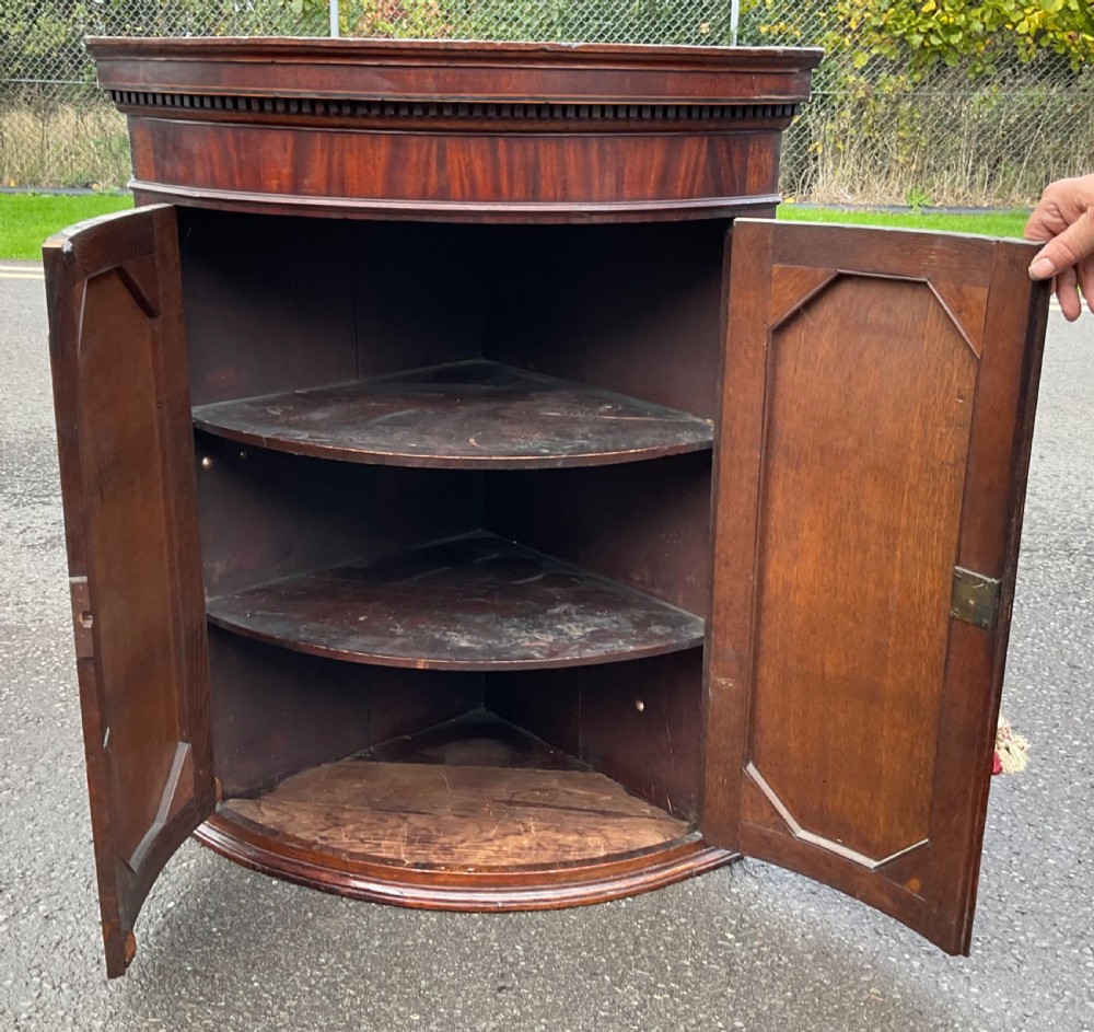 An antique Georgian mahogany bowfront hanging corner cupboard featuring twin doors with fan inlays and dentil moulding. View VI