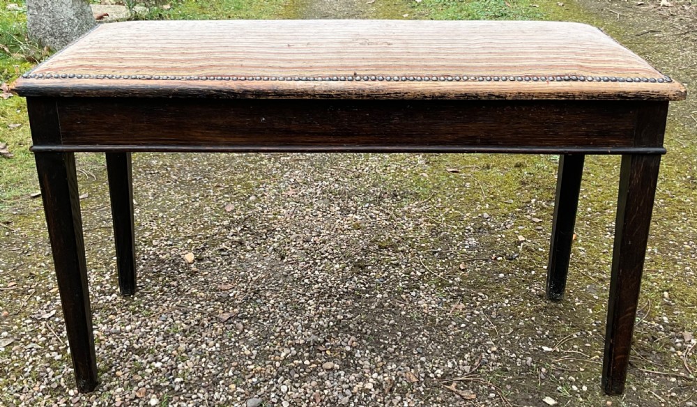 An Edwardian dark mahogany duet piano stool featuring four tapered legs and a padded striped fabric seat trimmed with brass studs.